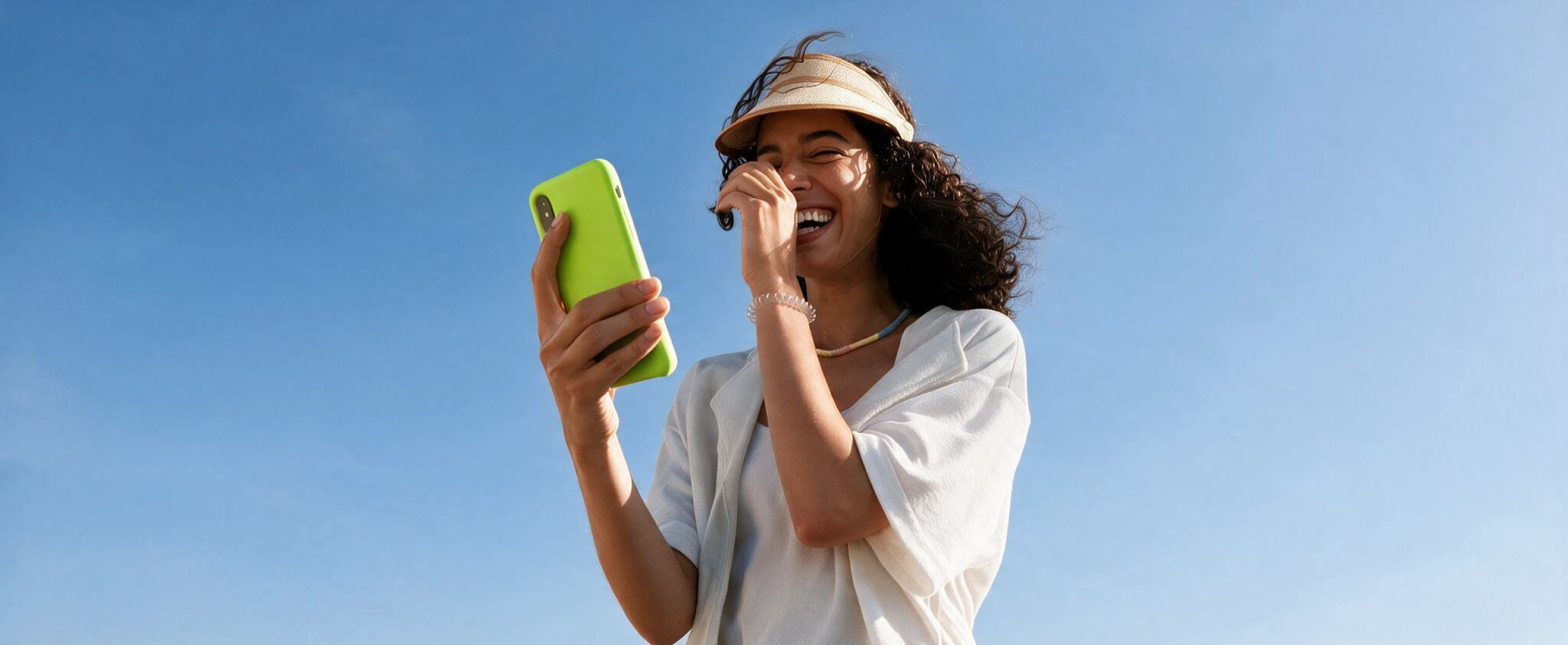 A woman laughing while looking at her phone against a clear blue sky